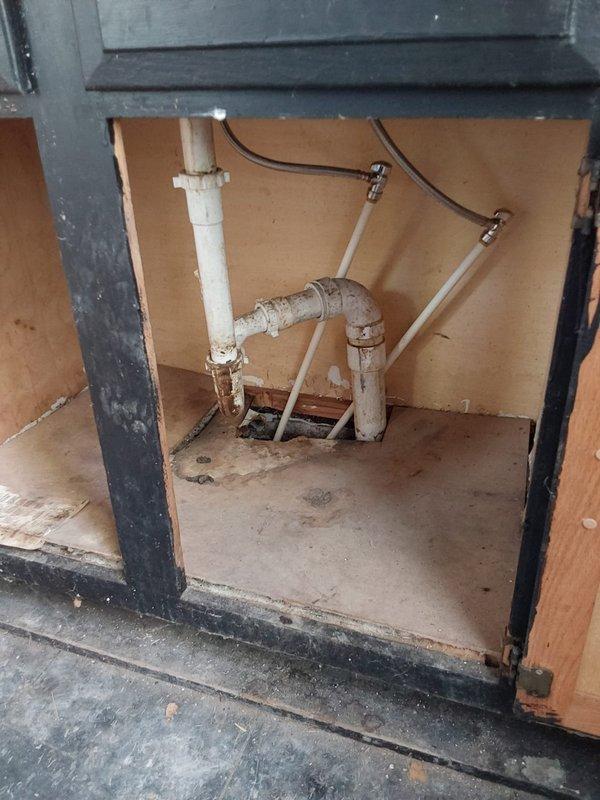Conducted comprehensive plumbing assessment for complete residential piping replacement. Property contains unused plumbing system requiring full replacement of all water supply and drainage lines throughout the home.

Inspected under-cabinet plumbing configuration including white PVC drainage assembly with P-trap, vertical and horizontal runs, and flexible supply lines secured with metal clamps. Noted significant age-related deterioration including discoloration, wear patterns, and accumulated debris on cabinet floor surfaces. Observed tan cardboard backing material and staining throughout accessible areas beneath counter spaces.

Evaluated exposed rough-in plumbing within floor cavity and wall framing systems. Documented multiple white PVC pipe runs converging at Y-junction fittings, vertical descending lines with elbow connections, and horizontal drainage runs between wooden joists. Identified dark-colored flexible components integrated within framing members alongside pink fiberglas