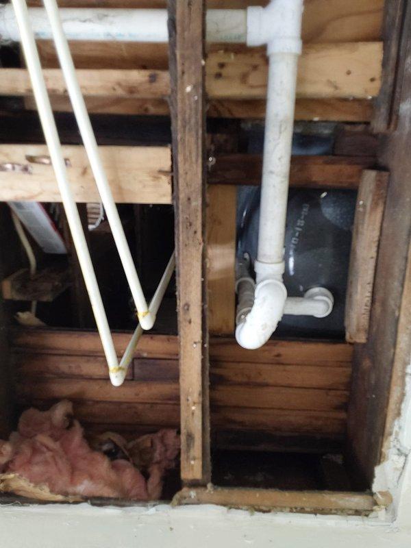 Conducted comprehensive plumbing assessment for complete residential piping replacement. Property contains unused plumbing system requiring full replacement of all water supply and drainage lines throughout the home.

Inspected under-cabinet plumbing configuration including white PVC drainage assembly with P-trap, vertical and horizontal runs, and flexible supply lines secured with metal clamps. Noted significant age-related deterioration including discoloration, wear patterns, and accumulated debris on cabinet floor surfaces. Observed tan cardboard backing material and staining throughout accessible areas beneath counter spaces.

Evaluated exposed rough-in plumbing within floor cavity and wall framing systems. Documented multiple white PVC pipe runs converging at Y-junction fittings, vertical descending lines with elbow connections, and horizontal drainage runs between wooden joists. Identified dark-colored flexible components integrated within framing members alongside pink fiberglas