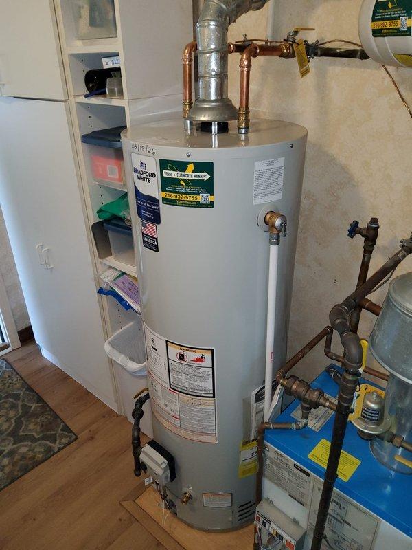Responded to service call for non-functional tanked water heater with reported gas leak and occasional "snowing" condition in 70-year-old residence. Upon arrival, inspected the white cylindrical water heater unit and surrounding plumbing connections, including copper piping, brass fittings, galvanized ductwork, and expansion tank assembly. Documented existing installation configuration showing multiple pipe connections at top of unit, adjacent blue water treatment system, and utility room conditions. Due to safety concerns regarding the reported gas leak, further diagnostics and corrective action were required to address both the operational failure and hazardous gas condition.