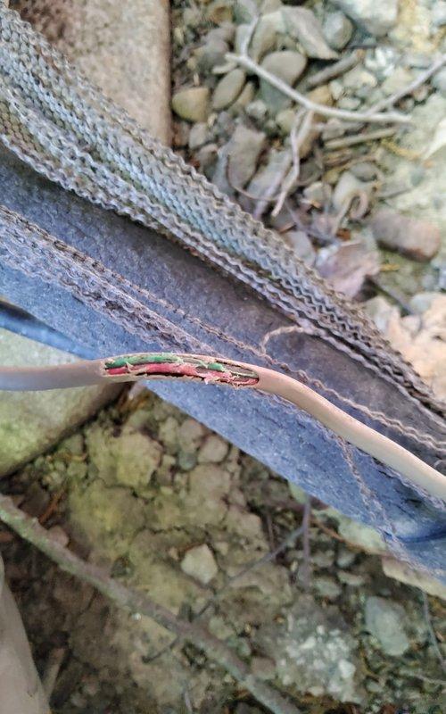 Responded to service call for 15-year-old Lennox air conditioning system. Upon arrival, performed visual inspection of outdoor condenser unit and surrounding components. Discovered damaged electrical wiring with exposed conductors near the unit, including compromised insulation jacket revealing internal green and red wires with exposed copper strands. The damaged wire was found lying across deteriorated cardboard material in the gravel base area, presenting a safety hazard and potential cause of system malfunction.

Inspected electrical disconnect box and conduit connections mounted on exterior brick wall. Assessed the condition of the cylindrical condenser unit with horizontal louvers and top-mounted fan grille. Documented the compromised wiring condition and deteriorated materials around the unit base that could impact electrical safety and system performance. 

Addressed the electrical hazard and restored safe operating conditions to the system. Verified proper connections at the di