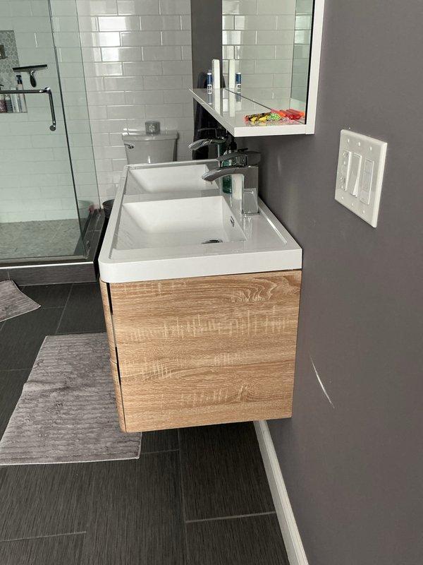 Completed plumbing estimate for new bathroom vanity installation featuring a wall-mounted double sink unit with wood-grain laminate cabinetry and white integrated countertop. Assessed rough-in plumbing connections, drain configuration for dual basins, and mounting requirements for the vanity cabinet, confirming compatibility with existing water supply lines and waste connections.