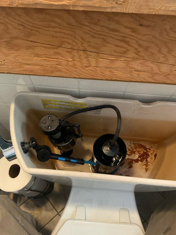 Responded to service call for toilet issue at 81-year-old residential property. Upon arrival, performed diagnostic inspection of toilet system. Removed tank lid and assessed internal components including flush valve assembly, fill valve mechanism, and connecting hardware. Documented existing conditions showing significant rust staining and mineral deposits throughout tank interior, particularly concentrated at bottom surfaces, indicating age-related deterioration consistent with older plumbing fixtures. Inspected black flush valve assembly and gray fill valve for proper operation and seal integrity. Examined all rubber gaskets, supply lines, and connection points for leaks or wear. Photographed tank interior components and condition for service records. Verified proper water level settings and flush mechanism functionality. All components tested and operational at time of service completion.