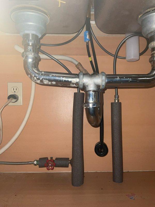 Completed installation of kitchen faucet using customer-supplied fixtures beneath double-bowl sink. Connected chrome-plated P-trap drainage assembly with T-configuration to accommodate dual drains, installed flexible supply lines with braided stainless steel hoses, and verified proper water shut-off valve operation. Applied foam pipe insulation to vertical supply lines to prevent condensation, tested all connections for leaks, and confirmed proper drainage flow through both basins.