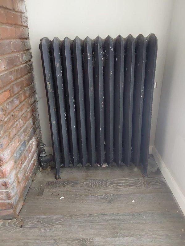 Performed radiator service and inspection in residential space. Updated lockbox access code to FAL per customer request via chat communication. Assessed traditional cast iron column-style radiator installation with approximately 10-11 sections positioned against interior wall. Verified proper valve and pipe connections at base of unit. Documented existing conditions including exposed brick wall adjacent to heating system and modern flooring installation. Noted minor surface wear and paint residue on radiator exterior consistent with age of equipment. Inspected window frame components and identified moisture-related deterioration with rust staining and finish degradation requiring monitoring. System operational upon completion of service call.