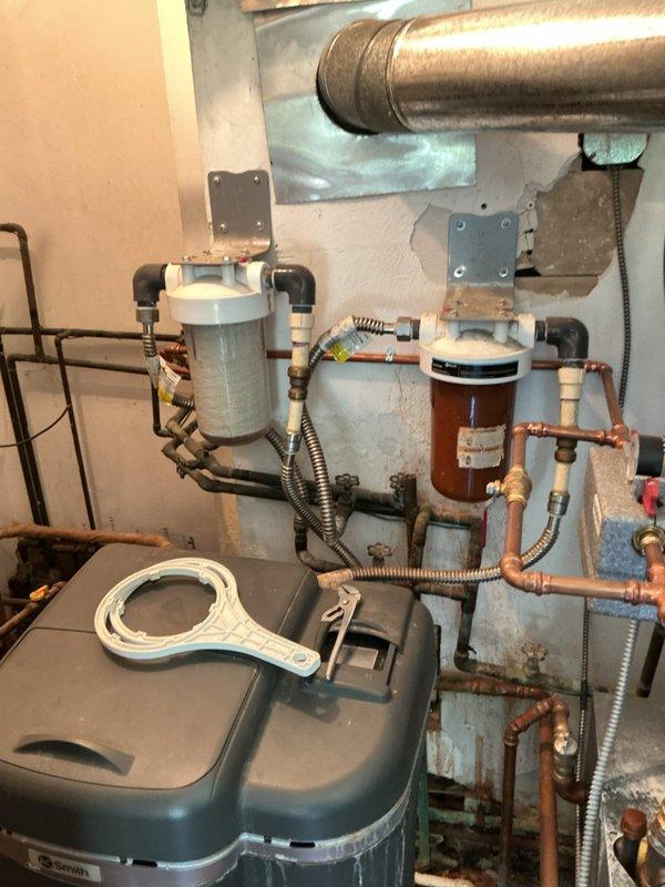 Responded to low water pressure complaint at 60-year-old residential property. Upon inspection of mechanical room, identified sediment buildup in dual water filter housings as primary cause of reduced flow. Removed and replaced both filter cartridges in white cylindrical housings connected to copper supply lines. Inspected water softener system for proper operation and verified brine tank levels were adequate. Checked pressure tank and expansion tank connections for proper function. Flushed sediment from copper piping system and tested pressure at multiple fixtures throughout home. Verified all valves in supply line were fully open and operating correctly. Restored water pressure to normal operating range. Recommended establishing regular filter replacement schedule every 3-6 months to prevent future pressure loss issues. System tested and functioning properly at completion.