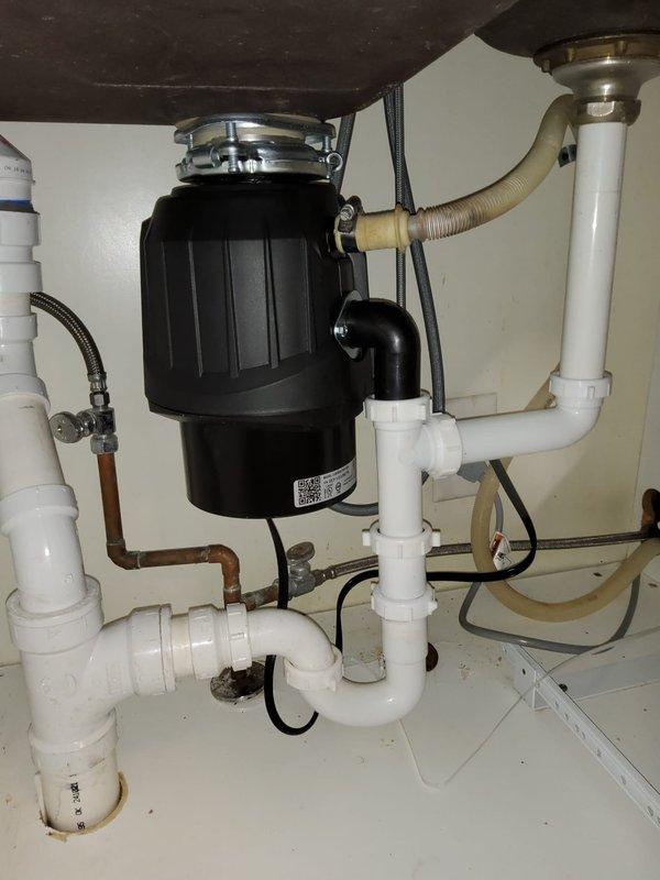 Responded to garbage disposal issue in century-old home. Upon arrival, inspected kitchen sink and under-sink plumbing configuration. Found black garbage disposal unit properly mounted to white rectangular single-basin sink with chrome faucet and standard PVC drainage assembly. Verified disposal mounting flange connection at sink drain, checked electrical wiring and power supply, and examined discharge plumbing including black elbow joint connecting to white PVC drain lines. Inspected dishwasher drain hose connection, water supply lines with braided flex connectors, and cleanout access point. Tested disposal operation and confirmed proper function of all components. System operating normally with no leaks detected at time of service completion.