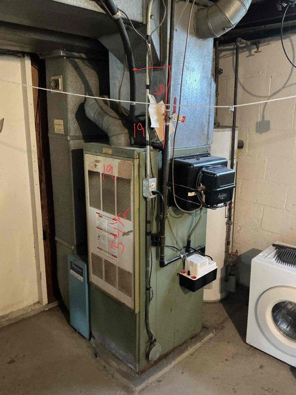 Completed on-site assessment for furnace replacement in residential basement utility room. Documented existing older model furnace unit with connected ductwork system, including sheet metal supply ducts, blue metal plenum, and wall-mounted humidifier or air treatment device. Photographed current HVAC configuration and mechanical room layout to prepare comprehensive replacement estimate for homeowner.