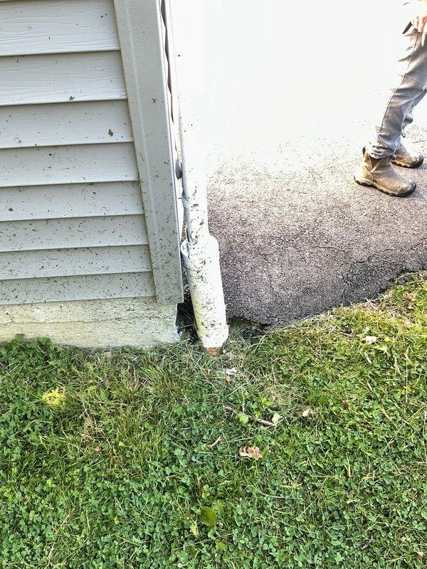 Provided estimate for extending downspout drainage system from building foundation to road. Assessment included evaluation of existing white vinyl downspout terminating at building base with inadequate drainage infrastructure. Current configuration showed downspout discharging directly onto concrete/paved surface adjacent to foundation, creating potential for water infiltration and foundation damage. Recommended installation of underground drainage piping to redirect roof runoff away from structure and convey water to road drainage system, ensuring proper grading and slope for gravity flow.