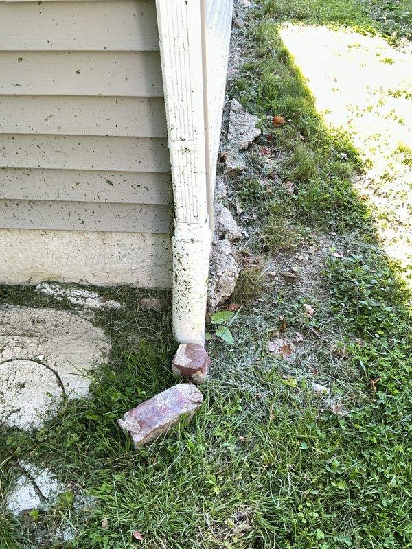 Provided estimate for extending downspout drainage system from building foundation to road. Assessment included evaluation of existing white vinyl downspout terminating at building base with inadequate drainage infrastructure. Current configuration showed downspout discharging directly onto concrete/paved surface adjacent to foundation, creating potential for water infiltration and foundation damage. Recommended installation of underground drainage piping to redirect roof runoff away from structure and convey water to road drainage system, ensuring proper grading and slope for gravity flow.