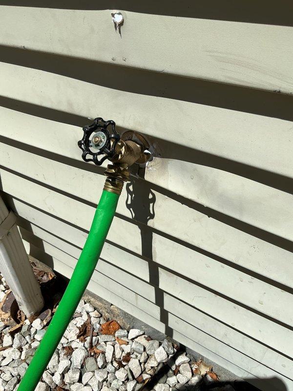 Responded to service call for severely leaking outdoor spigot. Accessed water supply line through interior wall panel to locate and address source of leak. Inspected copper supply piping, compression fittings, and brass shut-off valve with red handle located between floor joists. Evaluated exterior hose bib mounted on building siding and verified connection integrity where green garden hose was attached. Completed necessary repairs to eliminate water leak and restore proper function to outdoor water spigot system.