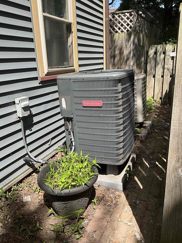 Conducted on-site evaluation for heat pump replacement at residential property. Assessed existing Goodman outdoor condenser units positioned along exterior wall with black and white striped siding. Documented current equipment condition, noting weathered appearance, dirt accumulation, and discoloration on existing unit. Evaluated installation site conditions including concrete pad placement, electrical conduit routing, clearances, and surrounding landscape features. Provided comprehensive replacement estimate for new heat pump system installation.