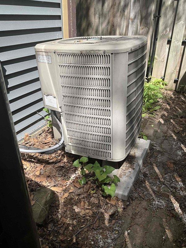 Conducted on-site evaluation for heat pump replacement at residential property. Assessed existing Goodman outdoor condenser units positioned along exterior wall with black and white striped siding. Documented current equipment condition, noting weathered appearance, dirt accumulation, and discoloration on existing unit. Evaluated installation site conditions including concrete pad placement, electrical conduit routing, clearances, and surrounding landscape features. Provided comprehensive replacement estimate for new heat pump system installation.