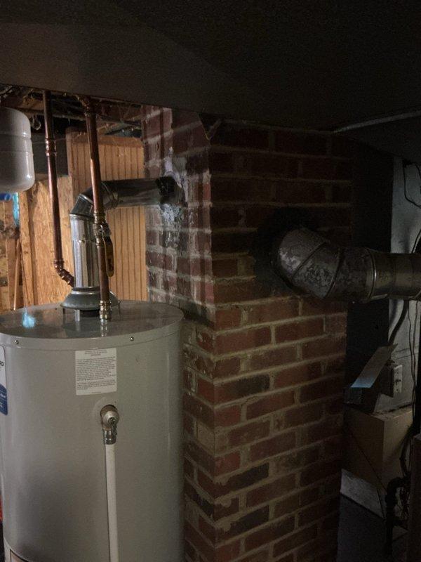 Conducted furnace replacement estimate for 22-year-old heating system located in basement mechanical room. Evaluated existing gray furnace unit positioned against concrete block wall, assessed ductwork configuration including large metal supply ducts, and documented current installation including proximity to masonry chimney structure and water heater. Photographed equipment specifications, warning labels, and surrounding utility infrastructure to provide comprehensive replacement recommendations.
