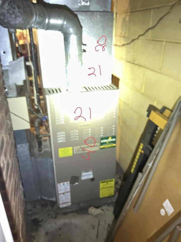 Conducted furnace replacement estimate for 22-year-old heating system located in basement mechanical room. Evaluated existing gray furnace unit positioned against concrete block wall, assessed ductwork configuration including large metal supply ducts, and documented current installation including proximity to masonry chimney structure and water heater. Photographed equipment specifications, warning labels, and surrounding utility infrastructure to provide comprehensive replacement recommendations.