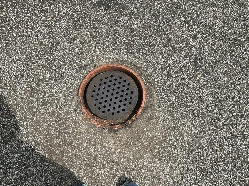 # Driveway Drain Repair and Cleaning

Responded to service call for damaged outdoor driveway drain at century-old residential property. Upon arrival, assessed crushed drain assembly that had been compromised by heavy vehicle traffic. The existing drain consisted of a perforated metal grate set within a copper/bronze collar frame, showing significant structural damage and displacement from its original position.

Removed damaged grate and inspected drain opening, which revealed a terracotta or ceramic collar with considerable weathering consistent with the property's age. Interior examination showed substantial accumulation of organic debris, decomposed leaves, and sediment buildup restricting proper drainage flow. The existing grate displayed advanced corrosion, rust damage, and mineral deposits throughout the perforated surface.

Cleared all debris and organic matter from drain opening and underlying pipe to restore proper water flow capacity. Cleaned terracotta collar and surrounding