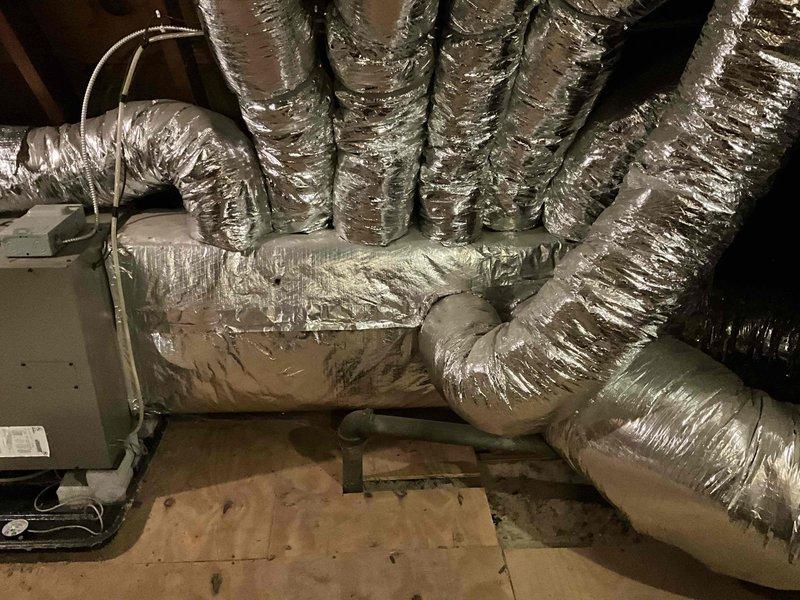 Conducted comprehensive HVAC system assessment in attic space for complete system replacement estimate. Evaluated existing air handler unit with associated flexible metallic ductwork distribution network throughout the residential attic installation. Documented current equipment configuration including beige air handler mounted on drain pan with white PVC condensate drainage piping and multiple runs of insulated flex duct extending to various zones. Assessed condition of existing ductwork showing typical wear patterns, compression points, and routing through confined attic space with exposed wooden roof trusses. Photographed complete system layout including air handler positioning, duct connections, support strapping, and overall installation accessibility for replacement planning purposes.