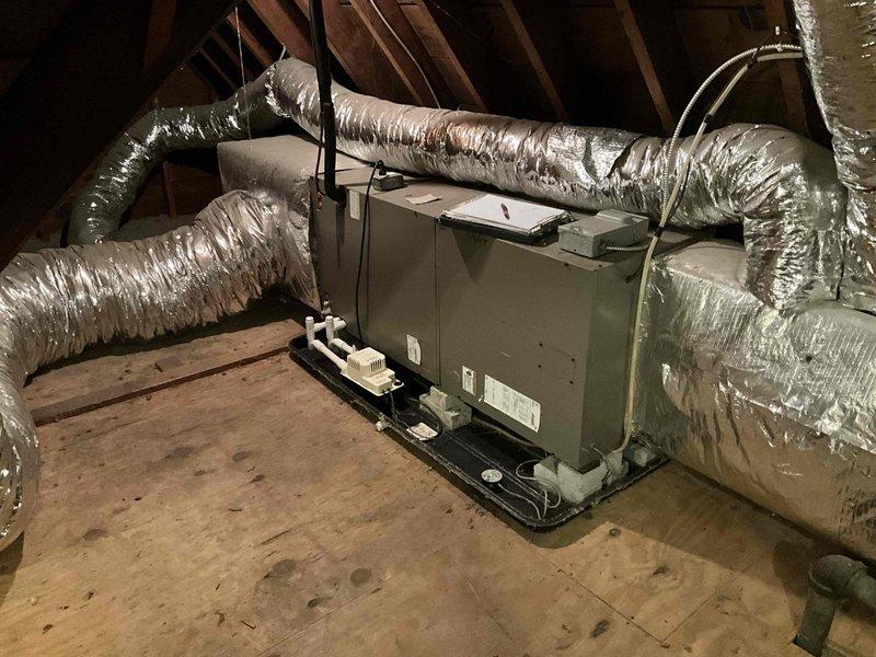 Conducted comprehensive HVAC system assessment in attic space for complete system replacement estimate. Evaluated existing air handler unit with associated flexible metallic ductwork distribution network throughout the residential attic installation. Documented current equipment configuration including beige air handler mounted on drain pan with white PVC condensate drainage piping and multiple runs of insulated flex duct extending to various zones. Assessed condition of existing ductwork showing typical wear patterns, compression points, and routing through confined attic space with exposed wooden roof trusses. Photographed complete system layout including air handler positioning, duct connections, support strapping, and overall installation accessibility for replacement planning purposes.