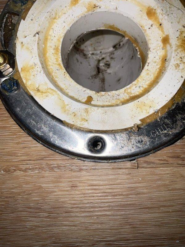Responded to service call regarding unstable toilet with urine seepage following previous stack repair. Upon inspection, discovered severely deteriorated toilet flange with extensive water damage and mineral staining, indicating prolonged leakage. The flange lacked proper structural support due to compromised subfloor material with no secure attachment point for mounting bolts. Removed damaged flange assembly and assessed PVC drain stack within wall cavity, which showed signs of previous repair work. Evaluated surrounding wooden framing and identified significant water damage to structural members, including weathered and discolored lumber requiring remediation. Documented existing conditions showing degraded wax ring gasket, stained flange surfaces, and compromised mounting area. Addressed structural deficiencies to provide stable base for proper flange installation and secure toilet mounting.