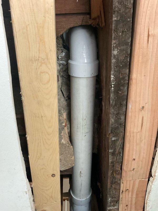 Responded to service call regarding unstable toilet with urine seepage following previous stack repair. Upon inspection, discovered severely deteriorated toilet flange with extensive water damage and mineral staining, indicating prolonged leakage. The flange lacked proper structural support due to compromised subfloor material with no secure attachment point for mounting bolts. Removed damaged flange assembly and assessed PVC drain stack within wall cavity, which showed signs of previous repair work. Evaluated surrounding wooden framing and identified significant water damage to structural members, including weathered and discolored lumber requiring remediation. Documented existing conditions showing degraded wax ring gasket, stained flange surfaces, and compromised mounting area. Addressed structural deficiencies to provide stable base for proper flange installation and secure toilet mounting.