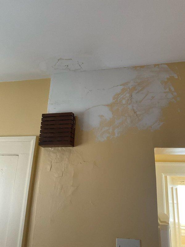 Responded to water leak emergency with visible ceiling and wall damage in room below bathroom. Upon inspection, identified active water intrusion causing significant discoloration, staining, and paint deterioration on ceiling and upper wall surfaces near ventilation grate. Traced source of leak to bathroom directly above affected area. Documented extensive water damage including ceiling sagging, wall staining with brownish discoloration, paint peeling, and exposed underlayer in multiple locations. Photographed damage to ceiling, walls, and bathroom fixtures for assessment and insurance documentation purposes.
