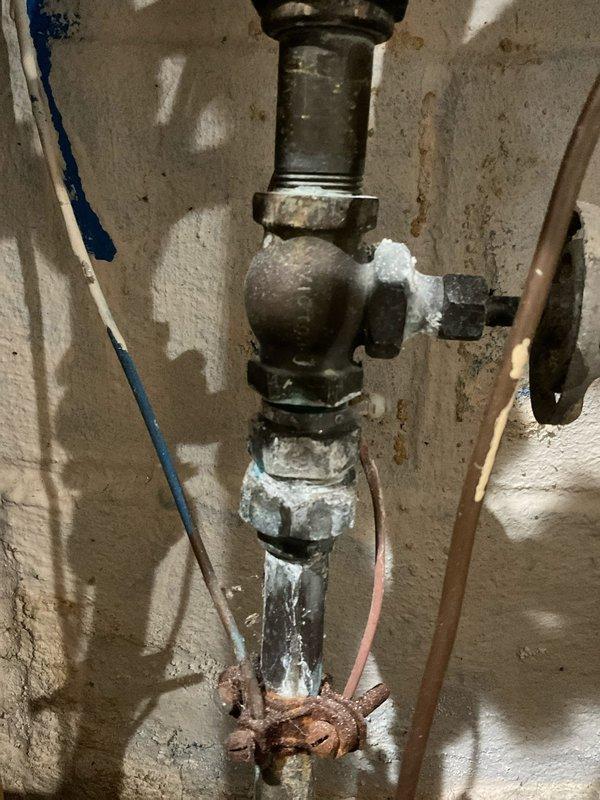 Responded to a service call for a clogged kitchen sink and leaking main water line shut off valve. Upon inspection, found the main shut off valve assembly showing severe corrosion, mineral buildup, and active leaking at threaded connections. The valve body and lower pipe sections exhibited extensive rust deterioration and white chalky mineral deposits indicating long-term water exposure and age-related failure. Documented the condition of the deteriorated plumbing assembly with photographs showing the corroded galvanized and brass fittings, compromised threaded connections, and oxidized components throughout the vertical pipe assembly.