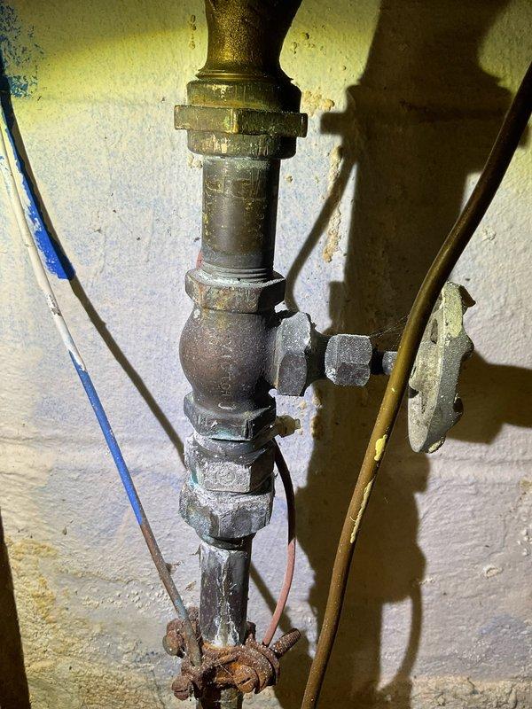 Responded to a service call for a clogged kitchen sink and leaking main water line shut off valve. Upon inspection, found the main shut off valve assembly showing severe corrosion, mineral buildup, and active leaking at threaded connections. The valve body and lower pipe sections exhibited extensive rust deterioration and white chalky mineral deposits indicating long-term water exposure and age-related failure. Documented the condition of the deteriorated plumbing assembly with photographs showing the corroded galvanized and brass fittings, compromised threaded connections, and oxidized components throughout the vertical pipe assembly.