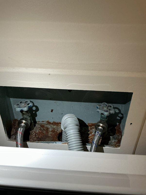 Responded to service call for leaking washing machine valve. Upon arrival, accessed the recessed plumbing panel and performed diagnostic inspection of the water supply connections. Identified active leak at one of the washing machine shut-off valves. Inspected both hot and cold water supply valves with cross-handle controls and the braided stainless steel supply lines. Noted significant rust and corrosion on the metal backing plate within the access panel, indicating prolonged moisture exposure from the leaking valve. Documented existing conditions with photographs showing the extent of corrosion damage and current plumbing configuration including supply valves, flexible supply lines, and corrugated drain hose installation.