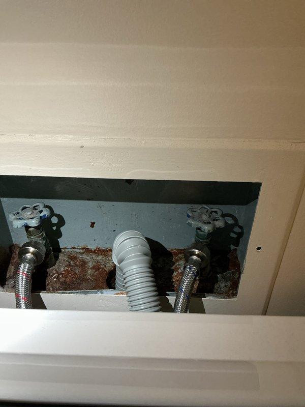 Responded to service call for leaking washing machine valve. Upon arrival, accessed the recessed plumbing panel and performed diagnostic inspection of the water supply connections. Identified active leak at one of the washing machine shut-off valves. Inspected both hot and cold water supply valves with cross-handle controls and the braided stainless steel supply lines. Noted significant rust and corrosion on the metal backing plate within the access panel, indicating prolonged moisture exposure from the leaking valve. Documented existing conditions with photographs showing the extent of corrosion damage and current plumbing configuration including supply valves, flexible supply lines, and corrugated drain hose installation.