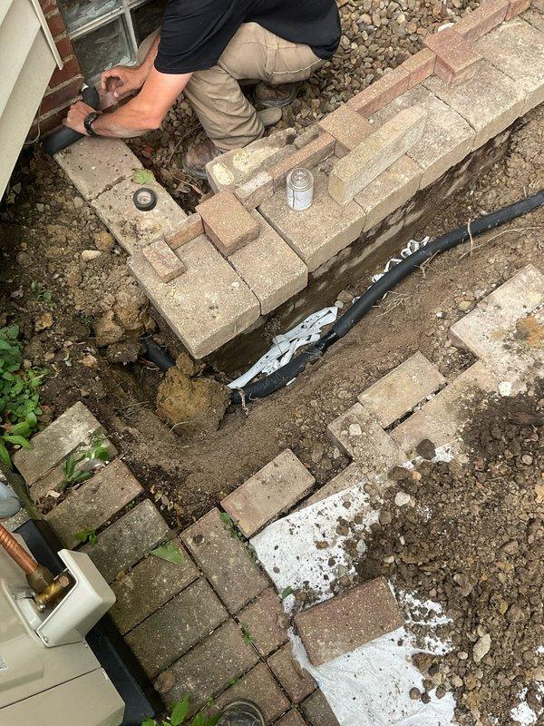 Removed and temporarily relocated concrete pavers in herringbone pattern to expose existing underground gas line path. Excavated and removed old gas line from interior of home through existing penetration near glass block window. Installed new grill stand and secured to ground with appropriate anchoring medium. Ran new underground gas line through original path, connecting from interior source point to new grill location. Assembled and installed outdoor grill unit, connected to new gas supply line, and performed complete leak test to verify system integrity and safe operation. Backfilled excavated areas, re-graded as necessary to ensure proper drainage around black corrugated conduit, and reset pavers over completed gas line installation with minimum 5-6 inch depth allowance for sand base and paver thickness.
