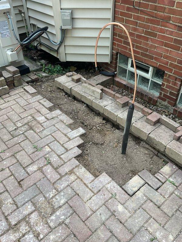 Removed and temporarily relocated concrete pavers in herringbone pattern to expose existing underground gas line path. Excavated and removed old gas line from interior of home through existing penetration near glass block window. Installed new grill stand and secured to ground with appropriate anchoring medium. Ran new underground gas line through original path, connecting from interior source point to new grill location. Assembled and installed outdoor grill unit, connected to new gas supply line, and performed complete leak test to verify system integrity and safe operation. Backfilled excavated areas, re-graded as necessary to ensure proper drainage around black corrugated conduit, and reset pavers over completed gas line installation with minimum 5-6 inch depth allowance for sand base and paver thickness.