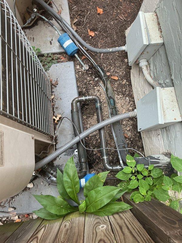 Conducted on-site evaluation for replacement of existing dual air conditioning system. Inspected two aging condenser units currently installed in side yard area, noting weathered condition, surface deterioration, and overall system age. Documented existing refrigerant line routing from condensers across concrete pad and through landscaped areas, including blue foam insulation sleeves on copper piping. Assessed electrical disconnect box, PVC conduit, and associated outdoor components. Photographed current installation configuration, equipment placement on ground surface, and surrounding fence/deck structures to determine optimal replacement approach. Provided comprehensive replacement estimate for new HVAC system installation.