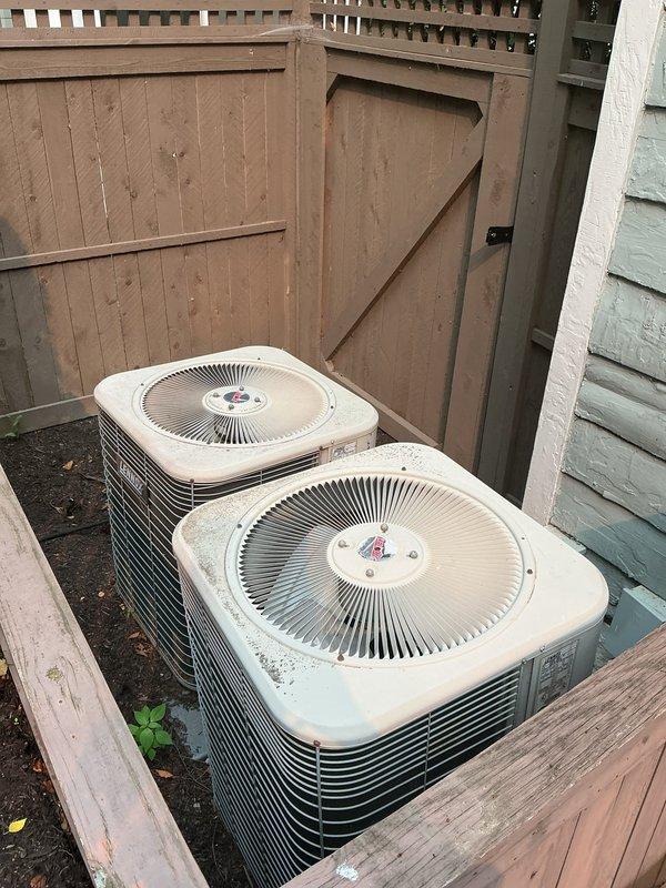Conducted on-site evaluation for replacement of existing dual air conditioning system. Inspected two aging condenser units currently installed in side yard area, noting weathered condition, surface deterioration, and overall system age. Documented existing refrigerant line routing from condensers across concrete pad and through landscaped areas, including blue foam insulation sleeves on copper piping. Assessed electrical disconnect box, PVC conduit, and associated outdoor components. Photographed current installation configuration, equipment placement on ground surface, and surrounding fence/deck structures to determine optimal replacement approach. Provided comprehensive replacement estimate for new HVAC system installation.