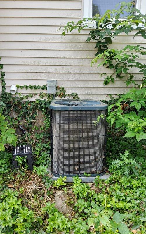 Performed second opinion evaluation on 15-year-old air conditioning system following previous contractor's diagnosis of refrigerant leak. Inspected outdoor condenser unit and found heavy vegetation overgrowth significantly restricting airflow around equipment, which can contribute to reduced efficiency and potential system strain. Verified thermostat operation showing 70°F room temperature setting in cooling mode. Documented excessive plant growth and debris accumulation around condenser requiring clearance for proper operation and serviceability.