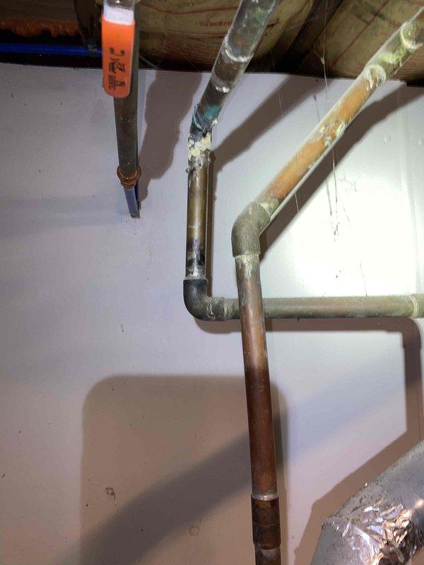 Responded to service call for reported boiler leak at 104-year-old residential property. Upon arrival, conducted thorough inspection of heating system and plumbing infrastructure in basement utility area. Examination revealed the concern was related to the water heater rather than the boiler system. 

Inspected white cylindrical water heater unit and found significant water staining and mineral deposits on top surface of tank, indicating previous moisture issues. Left copper pipe connection showed advanced corrosion and oxidation with reddish-brown discoloration at base fitting, suggesting long-term exposure to moisture. Pressure relief valve assembly on right side examined and found to be intact with proper metallic discharge piping in place.

Traced copper supply lines along wall and ceiling joists to identify potential leak sources. Tagged affected piping section for reference and documentation. Noted varying conditions of copper piping throughout system, with some sections showing 