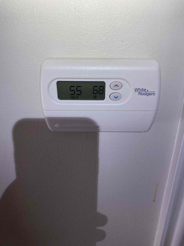 Responded to callback for thermostat malfunction following previous service visit. Inspected White-Rodgers digital thermostat displaying 55°F actual temperature and 68°F setpoint with system in hold mode. Removed thermostat cover and examined internal wiring connections, circuit board components, and AA battery power supply. Verified proper wire terminations at blue terminal blocks, checked for loose connections, and tested thermostat functionality to resolve ongoing temperature control issues.
