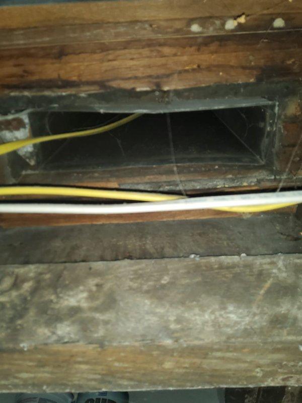 Responded to no-charge callback for assessment of insufficient airflow to back bedroom. Inspected ductwork in crawl space and attic area above affected room. Found rectangular duct opening in wooden floor framing with significant cobweb accumulation and signs of long-term neglect, indicating minimal to no air movement through this supply register. Documented exposed structural conditions, electrical wiring routing through duct space, and overall duct accessibility for future remediation work.