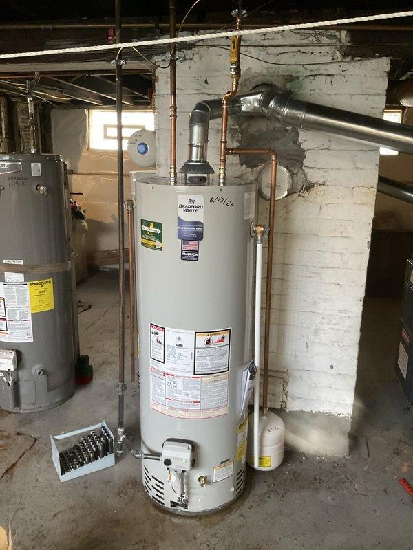 Replaced existing Bradford White residential water heater with new unit in basement utility area. Removed old water heater dated from 2014 and installed replacement gray cylindrical water heater. Connected copper supply and discharge piping to new unit with proper fittings and joints. Installed pressure relief valve with discharge pipe per code requirements. Connected metal exhaust vent pipe for proper combustion gas venting. Verified all plumbing connections for leaks and tested unit operation to ensure proper heating functionality.