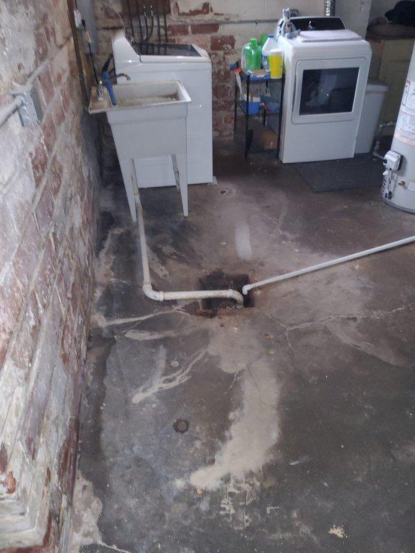 Responded to recurring main drain line backup causing basement flooding. Upon arrival, standing water had receded. Customer reported repeated backups with suspected root intrusion in main sewer line. Assessed basement area showing evidence of previous water damage on concrete floor with visible staining and moisture patterns. Located main drain cleanout access point with white PVC pipe and cleanout fitting on basement wall. Documented existing conditions including floor drain and plumbing configuration. Based on recurring nature of backups and customer history of blockages, recommended scheduling follow-up service with specialized equipment and additional technician for comprehensive main line cleaning and video camera inspection to confirm root intrusion and determine extent of blockage in sewer line.