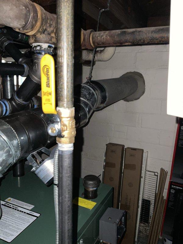 Responded to gas leak call at century-old residential property. Upon arrival, conducted thorough inspection of plumbing and gas piping system in basement mechanical room. Located active gas leak at valve assembly connected to aging gas piping infrastructure. White painted concrete block walls and corroded metal piping indicated extensive age of existing system components. 

Identified leak source at gas valve fitting near green cylindrical pressure tank with multiple pipe connections. Performed leak detection procedures and confirmed gas escape at metallic valve assembly. Tagged affected piping with identification marker for documentation purposes. Isolated gas supply to affected section and performed necessary repairs to valve connections and fittings. Tested all repaired connections with leak detection solution to verify complete seal integrity. Restored gas service after confirming no residual leaks present in system. 

Documented existing conditions including significantly corroded