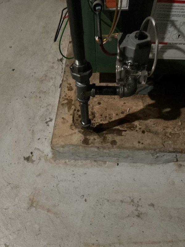 Responded to gas leak call at century-old residential property. Upon arrival, conducted thorough inspection of plumbing and gas piping system in basement mechanical room. Located active gas leak at valve assembly connected to aging gas piping infrastructure. White painted concrete block walls and corroded metal piping indicated extensive age of existing system components. 

Identified leak source at gas valve fitting near green cylindrical pressure tank with multiple pipe connections. Performed leak detection procedures and confirmed gas escape at metallic valve assembly. Tagged affected piping with identification marker for documentation purposes. Isolated gas supply to affected section and performed necessary repairs to valve connections and fittings. Tested all repaired connections with leak detection solution to verify complete seal integrity. Restored gas service after confirming no residual leaks present in system. 

Documented existing conditions including significantly corroded