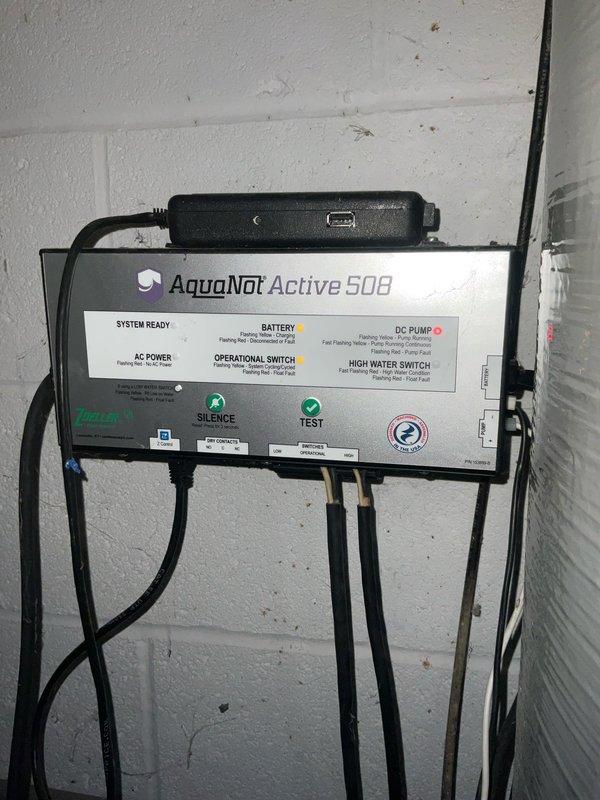 Responded to service call for AquaNot Active 500 battery backup sump pump system experiencing daily test mode failures with recurring alarm activation. System had been malfunctioning for several weeks, requiring customer to manually silence alarms. Inspected sump pump basin containing green primary pump unit with float switch assembly and associated PVC discharge piping. Evaluated AquaNot control panel mounted on wall, examining system ready indicators, battery status, DC pump operation, AC power supply, and high water switch functionality. Tested operational and silence buttons on control unit. Assessed battery backup component and electrical connections. Documented existing installation configuration including pump placement, plumbing connections, and control system setup through photographic evidence for service records.