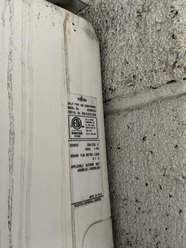 Completed HVAC estimate for replacement of existing air conditioning system in mechanical room. Evaluated Fujitsu split-type air conditioner condenser unit mounted on exterior brick wall with white concrete base. Unit operates on 200/230V electrical specifications and is connected via black electrical conduit running along building exterior. Documented equipment specifications, installation location, mounting configuration on metal brackets, and overall system condition. Photographed condenser unit, manufacturer specification label, electrical connections, and surrounding installation area to provide comprehensive assessment for replacement recommendation.