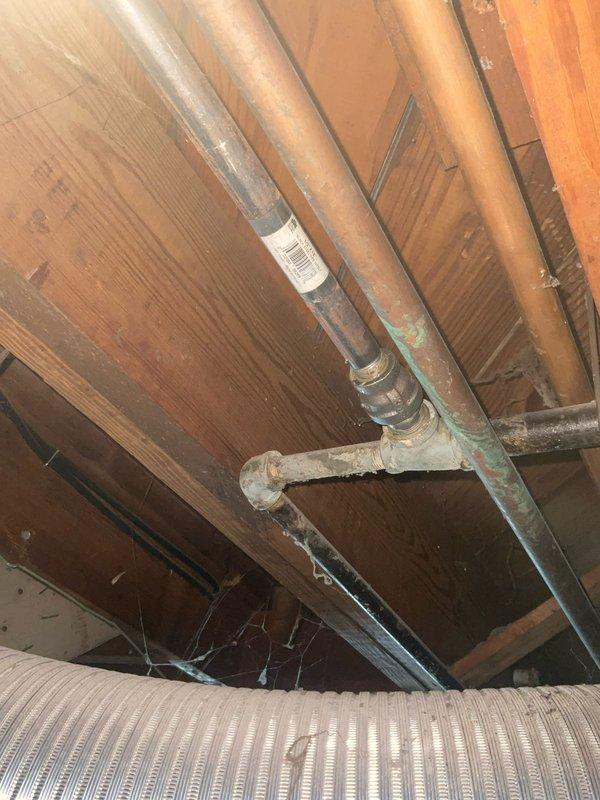 Performed comprehensive inspection of residential tanked water heater system at customer's request. Examined water heater installation in basement area, including all associated copper and PVC plumbing connections, pressure relief valve, and overhead piping distribution. Verified proper mounting and support of vertical discharge piping secured to ceiling joists. Inspected sump pump system mounted on white collection tank adjacent to water heater. Assessed condition of copper supply lines showing age-appropriate patina and verdigris oxidation. Documented all components with photographs for service records. System found to be operational with connections secure and properly supported.