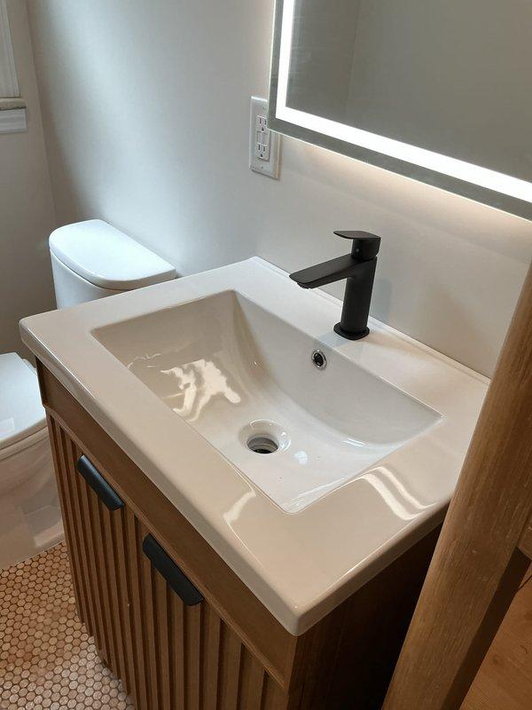 Completed installation of two toilets in separate bathroom locations. First bathroom featured white ceramic toilet installed in corner setting with existing subway tile wainscoting and basket-weave mosaic floor. Second bathroom installation included toilet positioning adjacent to new vanity setup. Set and secured vanity unit with light wood finish featuring vertical slat detailing, integrated white rectangular sink, and matte black single-handle faucet. Ensured proper connections, verified water supply lines, tested flush mechanisms and faucet operation for leaks, and confirmed all fixtures level and properly sealed.