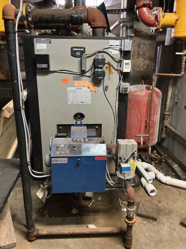 Conducted commercial boiler system assessment in mechanical room. Evaluated existing wall-mounted boiler unit with blue burner assembly, associated piping infrastructure including overhead orange supply lines, copper distribution piping, and aging red expansion tank. Documented current system configuration, control components, and ancillary equipment including yellow separator vessel and insulated ductwork for estimate preparation.
