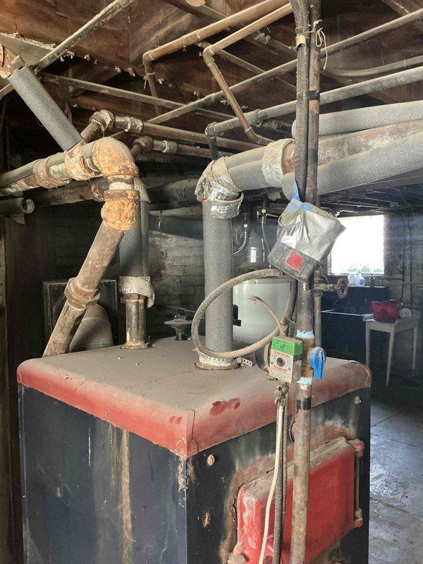 Conducted on-site evaluation of a century-old United States Radiator Company Redtop model boiler system for replacement estimate. The existing boiler exhibits significant aging with extensive corrosion and mineral buildup throughout the piping network, oxidized copper and brass fittings, and visible deterioration of the mechanical components. Documented current system condition through photographic assessment showing the main boiler cabinet, overhead piping infrastructure, and compromised pipe joints with heavy white mineral deposits indicating long-term moisture exposure and chemical degradation.