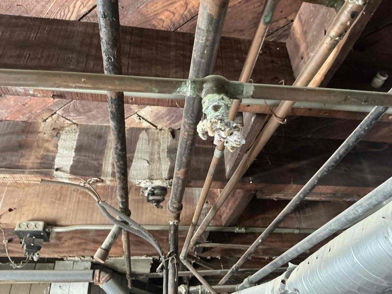 Conducted on-site evaluation of a century-old United States Radiator Company Redtop model boiler system for replacement estimate. The existing boiler exhibits significant aging with extensive corrosion and mineral buildup throughout the piping network, oxidized copper and brass fittings, and visible deterioration of the mechanical components. Documented current system condition through photographic assessment showing the main boiler cabinet, overhead piping infrastructure, and compromised pipe joints with heavy white mineral deposits indicating long-term moisture exposure and chemical degradation.
