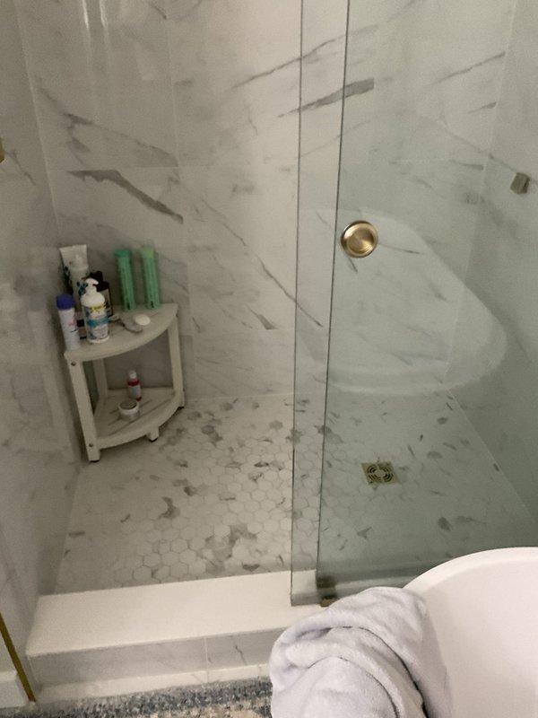 Conducted site assessment for bathroom plumbing upgrades. Evaluated existing water line routing to provide estimate for re-running supply line. Inspected toilet for removal and replacement, documenting current fixture condition and surrounding flooring. Assessed shower area for handicap grab bar installation, noting tile wall construction and optimal placement locations near tub/shower entry. Photographed all work areas including shower with marble-pattern tile walls and existing fixtures, toilet area with adjacent vanity, and surrounding bathroom infrastructure to support detailed estimate preparation for proposed plumbing modifications and accessibility improvements.