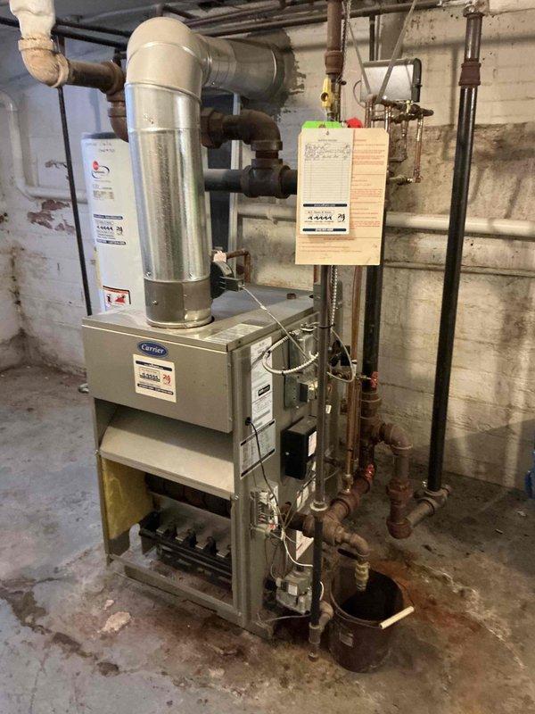 Conducted on-site assessment of existing Carrier heating system in basement mechanical room to provide steam boiler replacement estimate. Evaluated current equipment configuration, including furnace unit, ductwork, expansion tank, and associated plumbing connections. Documented existing installation with photographs showing multiple aged pipes with visible rust and corrosion on metal fittings, water-stained ceiling areas, and overall mechanical room layout. Assessed compatibility requirements for new steam boiler installation, including necessary pipe connections, venting requirements, and spatial considerations for equipment replacement in the existing footprint.