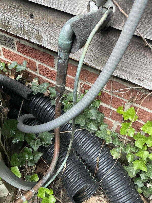 Conducted estimate for complete HVAC system replacement at residential property. Evaluated existing outdoor condenser unit located on side of building, which is surrounded by overgrown vegetation and shows signs of aging infrastructure including oxidized copper refrigerant lines and weathered electrical conduit. Documented current system condition with photographs for insurance claim purposes, noting the unit's accessibility issues due to dense plant growth and the deteriorated state of associated piping and utility connections at the building's foundation.